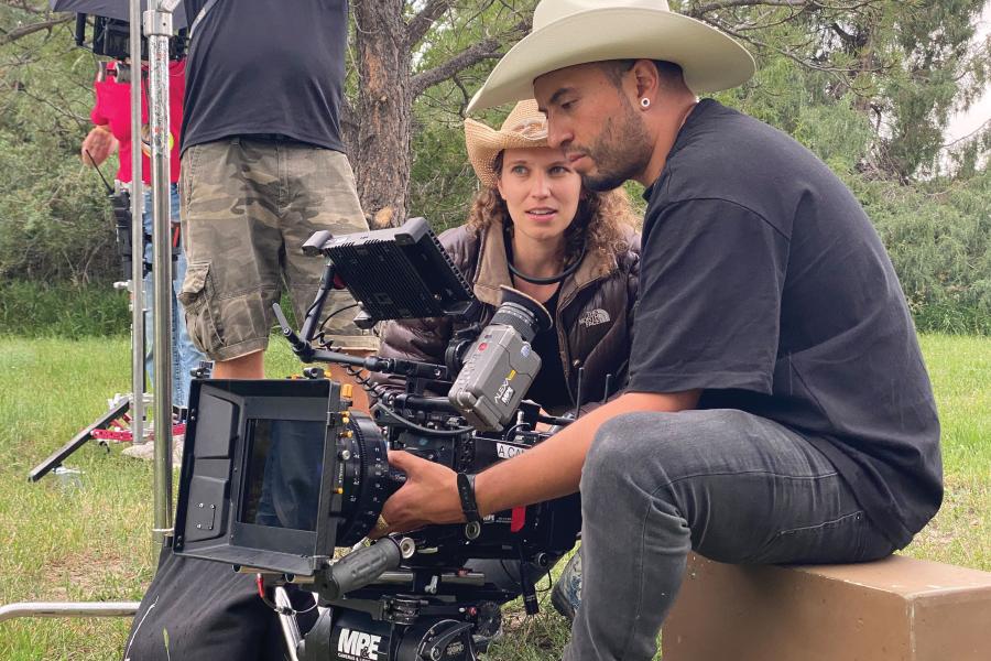 Emma Needell, wearing a cowboy hat, looks on as a someone holding a video camera films a scene