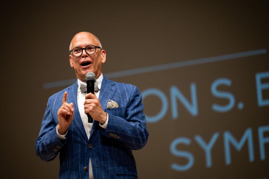 Jim Obergefell on stage at Johns Hopkins University; he hols a microphone in his left hand and is wearing a blue checked sports coat and a bowtie