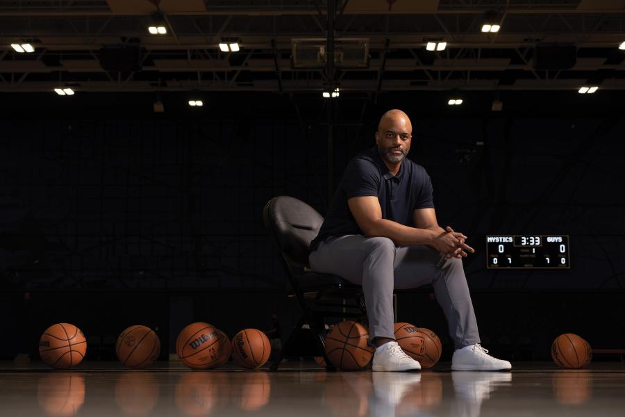 Wes Unseld Jr. sits on chair in an indoor basketball court. On the ground are several basketballs.