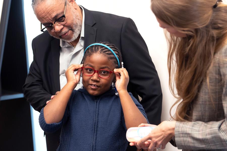 A young girl with a light blue headband tries on a pair of red eyeglasses 