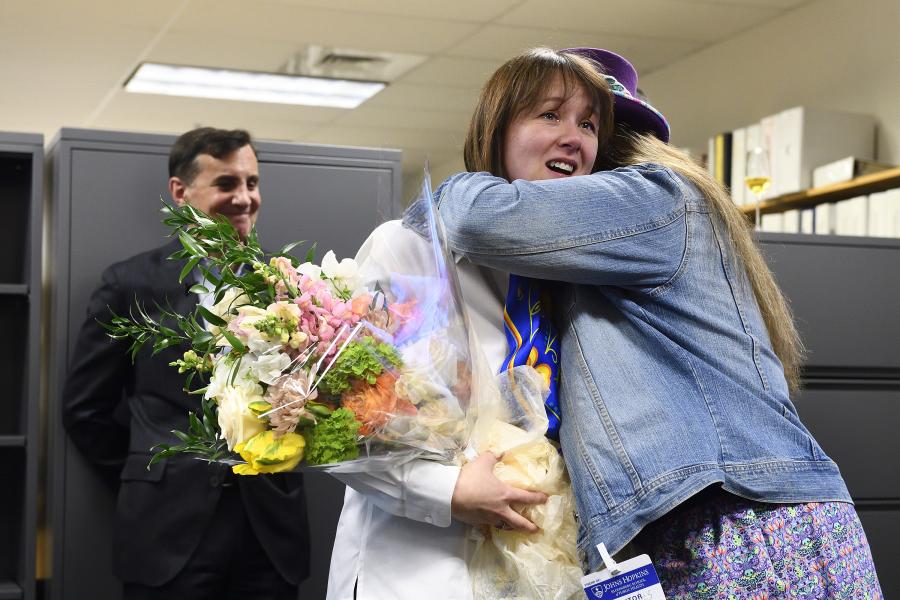 Melissa Walls is hugged by a colleague while holding a bouquet of white, yellow, orange, and pink flowers