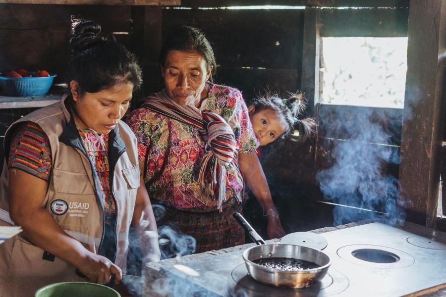 A Jhpiego/USAID worker stands next to an older woman, showing her how to cook a bean recipe. A toddler is strapped to the older woman's back.