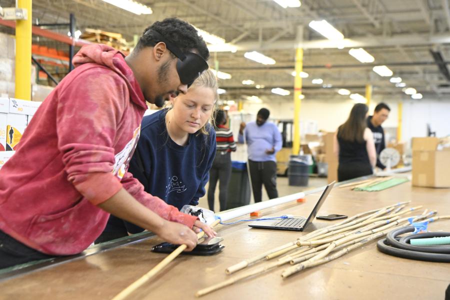 Students work in partnership with Blind Industries and Services of Maryland to develop a system that will allow blind individuals to manufacture canes. A student-associate pair works together at a long wooden work table.