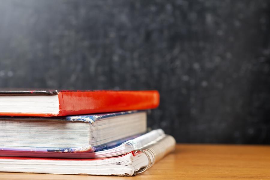 A stack of old textbooks on a wooden desk with a black chalkboard in the background
