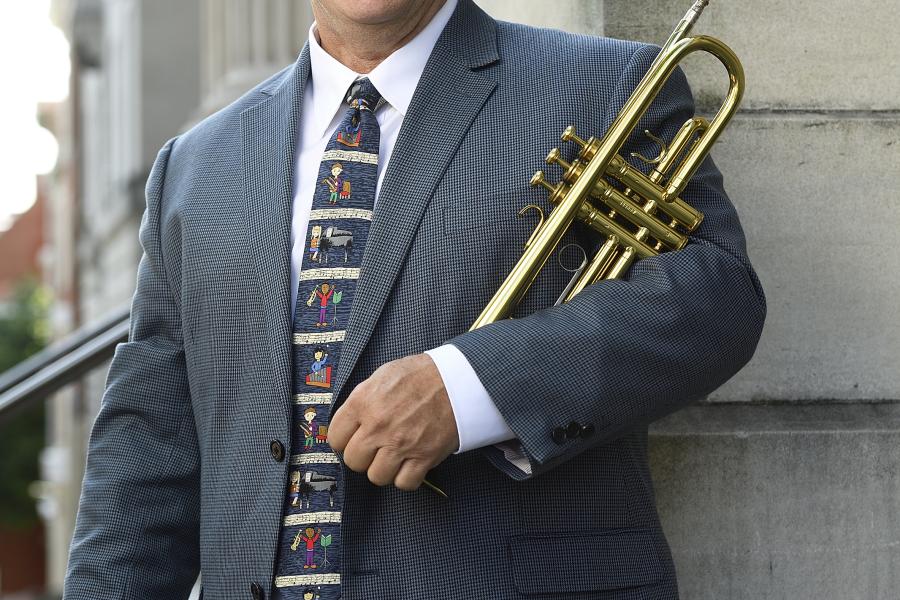 A man in a blue-gray sports coat and tie stands holding a trumpet