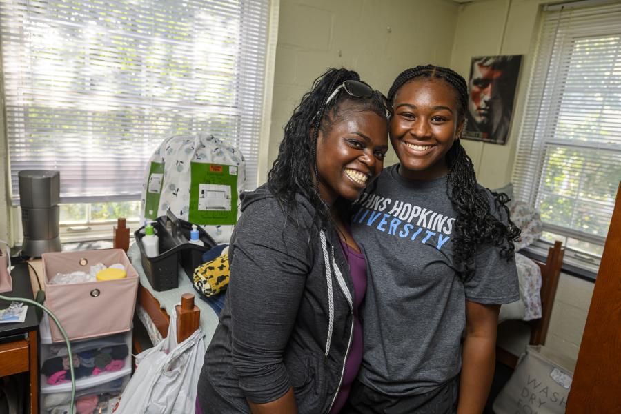 An incoming freshman and their parent smile for the camera. Behind, a dorm bed is covered with items from move-in.