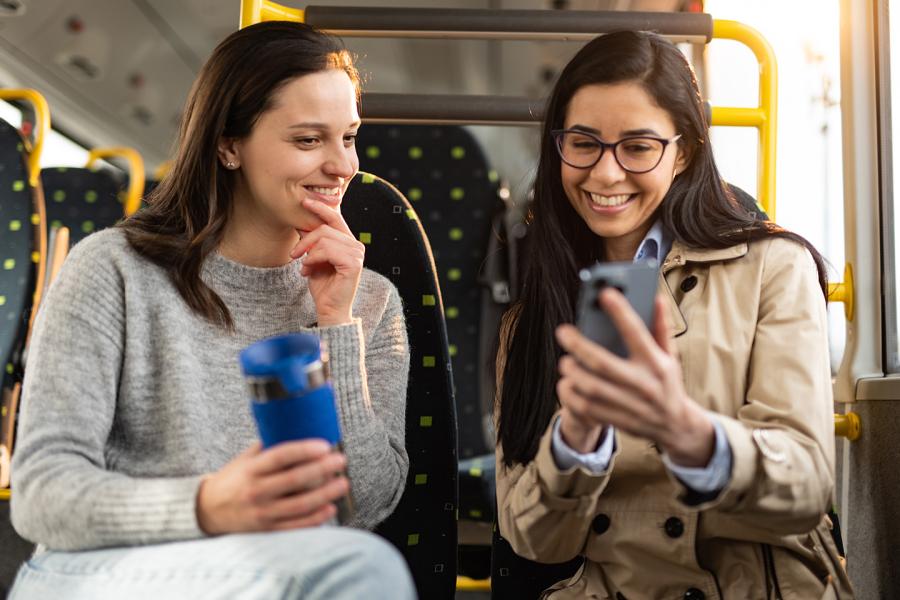 Two young women sitting next to each other on a bus look at something on one of their phones. The other person is holding a beverage cup. a 