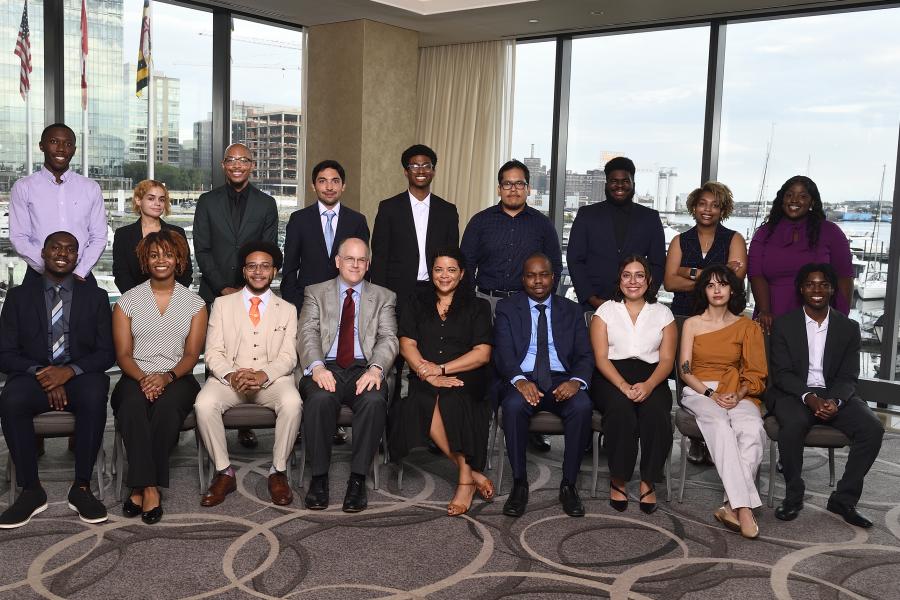 A group of 15 VTSI scholars plus three program leaders pose for a photo; they are arrange in two rows, with the front row sitting and the back row standing; boats and Baltimore's Inner Harbor can be seen through the large windows behind them