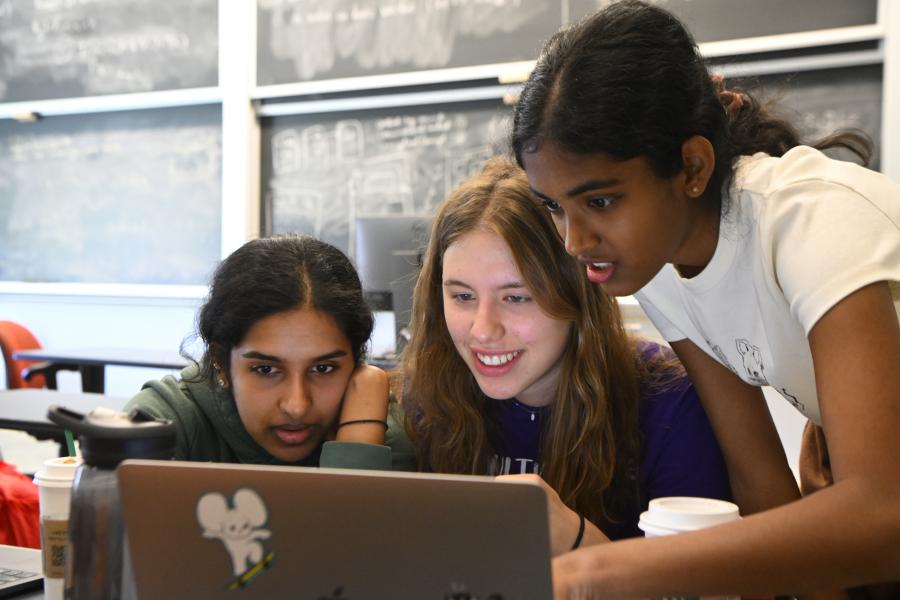 Three students lean in to look at a laptop