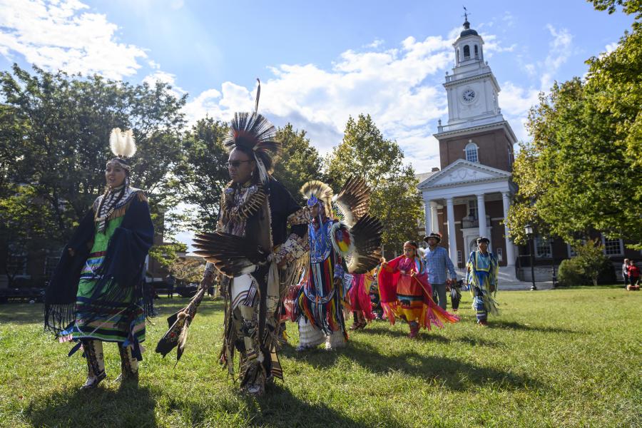 Indigenous men in traditional dress lead a line of people in dancing in front of Gilman Hall