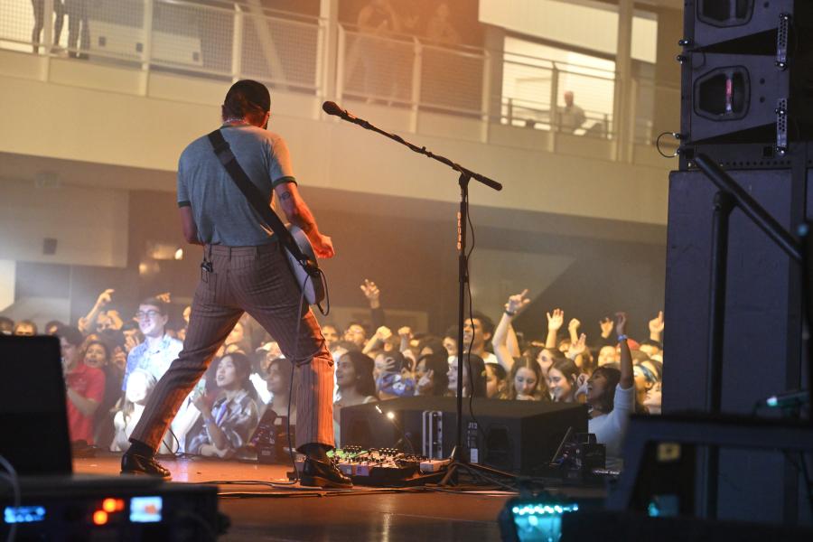 A musican plays an electric guitar onstage while facing a crowd, back to the camera