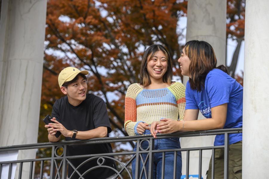 Three students lean against a railing while talking to each other.