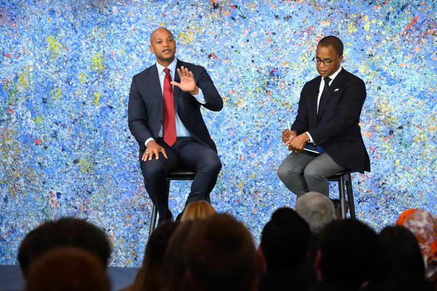 Two men sit on a stage in front of a multicolored art piece, addressing a large audience