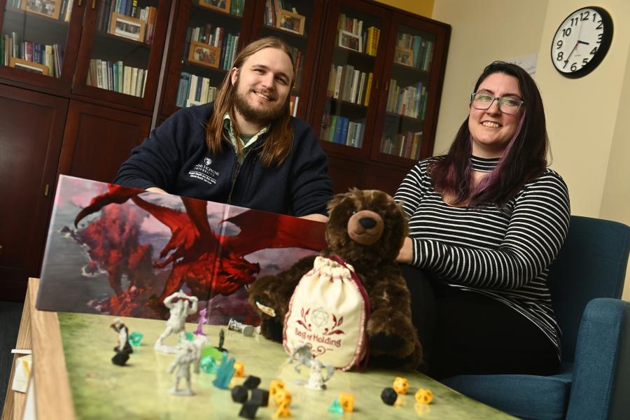 Two adults smile for the camera while sitting behind a table covered in Dungeons and Dragons pieces.