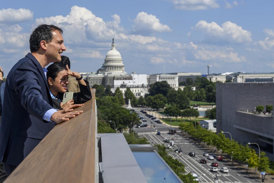 Two adults look out from a rooftop. Behind them is the US Capitol building.