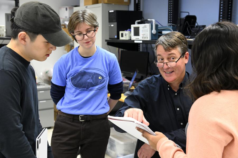 A professor explains something to three students, pointing at one of their notebooks