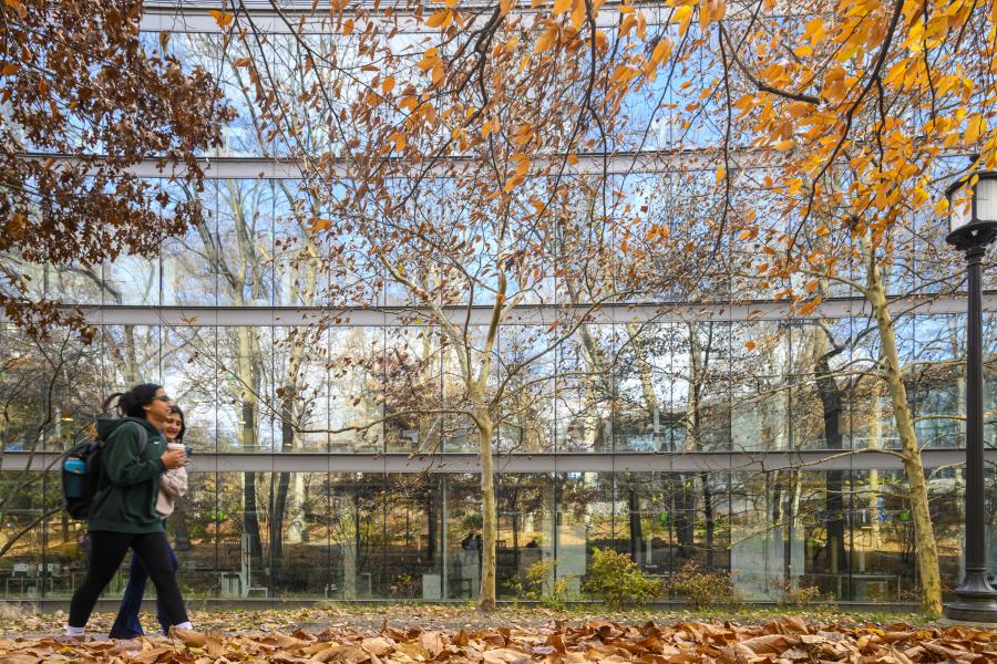 Two students walk past a glass building. The building is reflecting autumn trees.