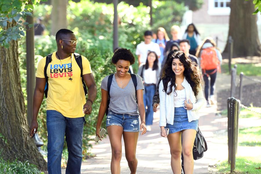 Three students walk along a brick pathway in a college campus setting; a larger group of students is visible in the background