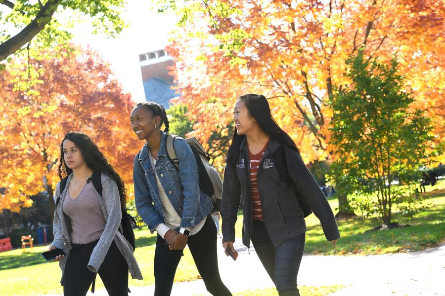 Three students walk across campus amidst orange and yellow fall foliage
