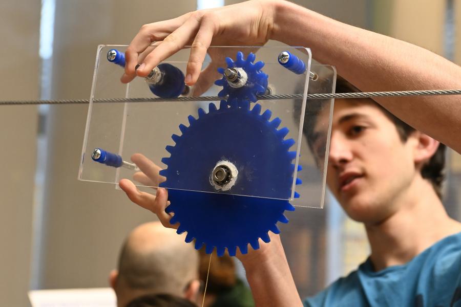A student balances a contraption made of gears on a wire.