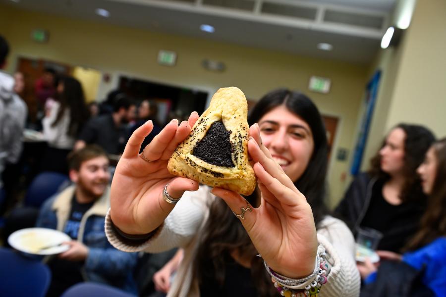 A female student smiles while holding a hamantaschen close to the camera