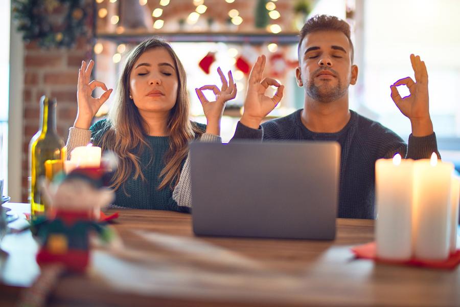 Sitting in a room filled with holiday decorations, a man and woman are meditating in front of an open laptop.