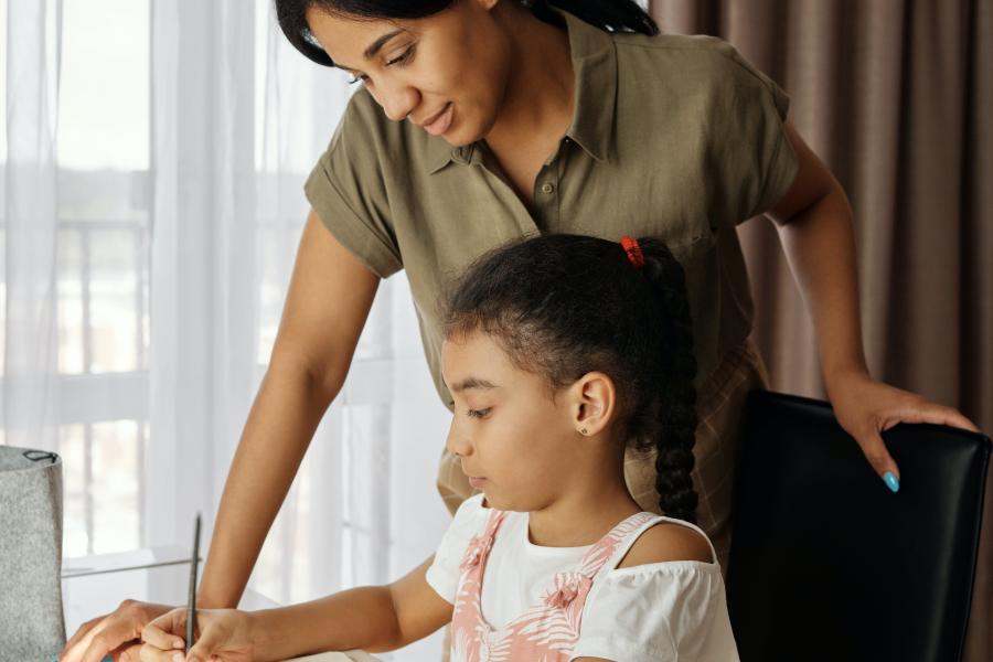 A daughter sits at a desk doing homework while her mom stands beside her helping