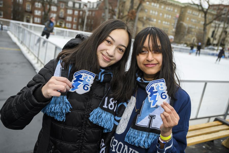 Two students stand in front of an ice rink, holding up stickers that read 