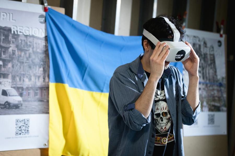 A man wears a VR headset while standing in front of a blue and yellow Ukraine flag