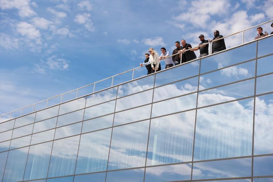 A group of people stand on the roof of a glass building. The building reflects the blue sky and clouds.