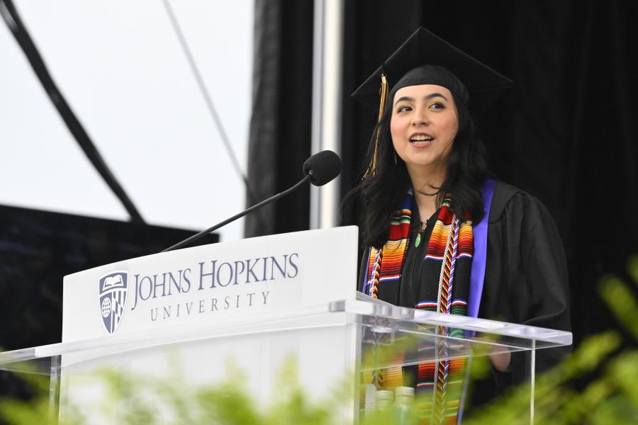 A student in a cap and gown speaks at a podium at JHU Commencement.