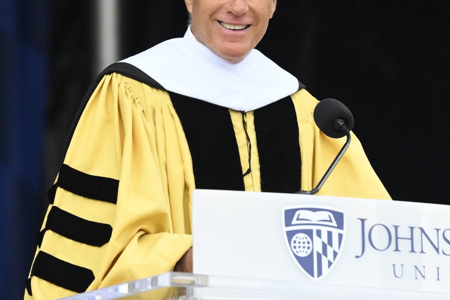 Mitt Romney wears yellow graduation regalia while standing behind a Johns Hopkins University podium.