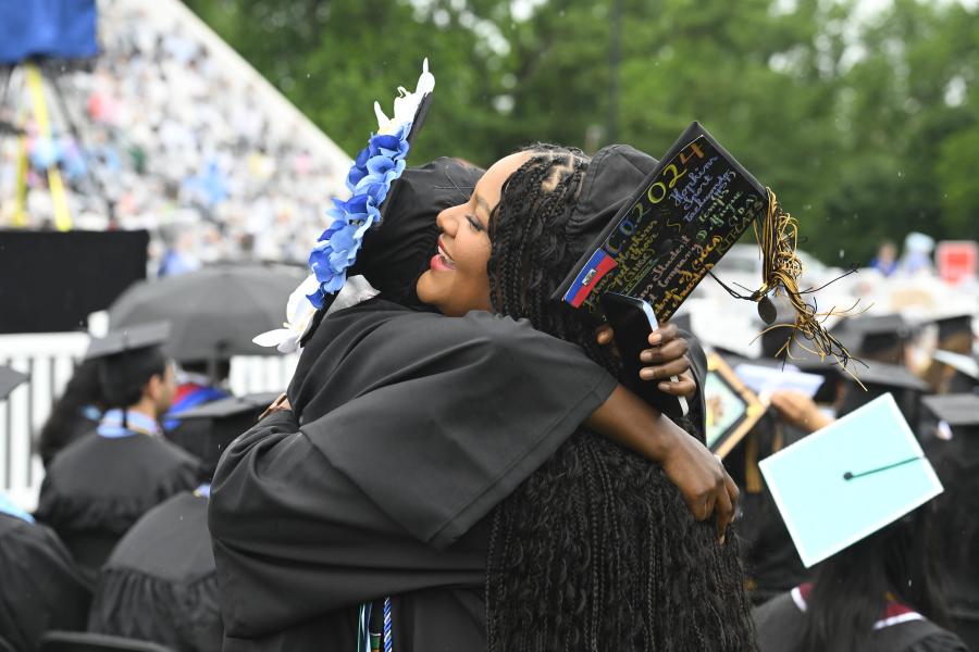 Two graduating students wearing caps and gowns hug each other.