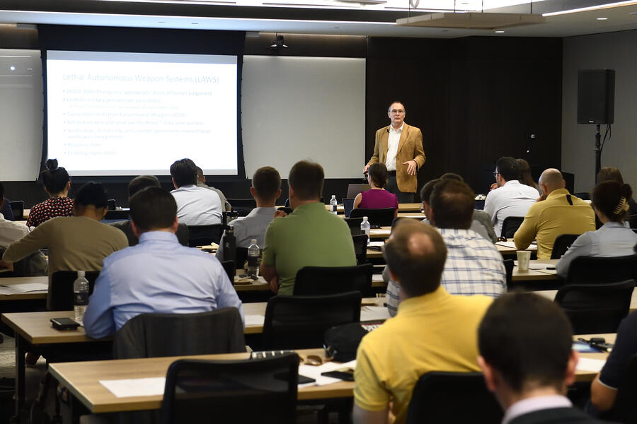 Jim Bellingham, an older white male, stands in front of a projector and addresses a crowded room