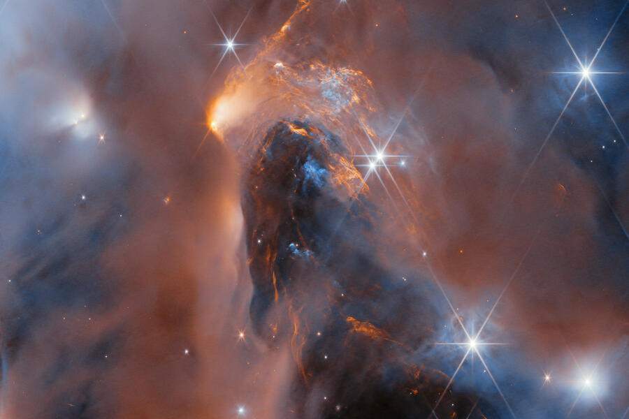 A nebula made up of cloudy gas and dust in the form of soft and wispy clouds and, in the centre, thin and highly detailed layers pressed close together. Large, bright stars surrounded by six long points of light are dotted over the image, as well as some small, point-like stars embedded in the clouds. The clouds are lit up in blue close to the stars; orange colours show clouds that glow in infrared light.