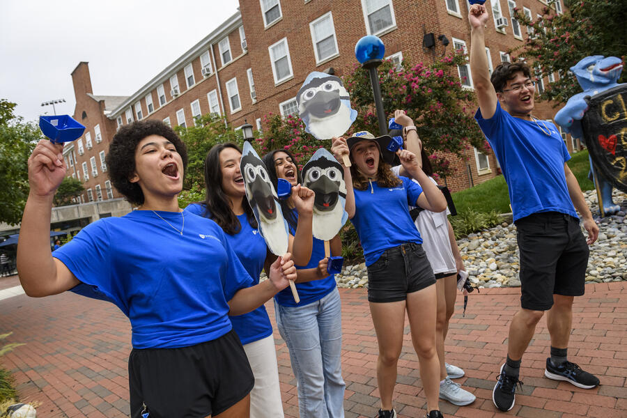 Six college students cheer while waving bells and headshots of Jay the Blue Jay during new student move-in day.