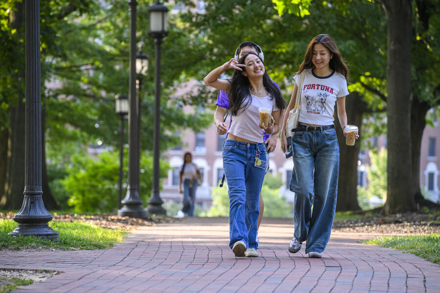 Two college students walk to class on a sunny day. One is making a peace sign for the camera.