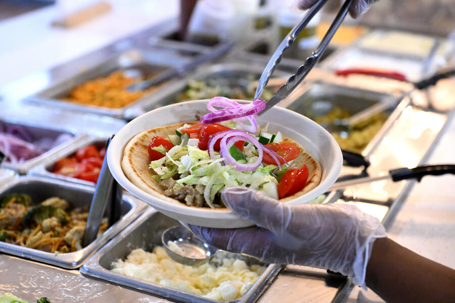 Gloved hands assemble a gyro in a paper bowl over a prep station full of ingredients.