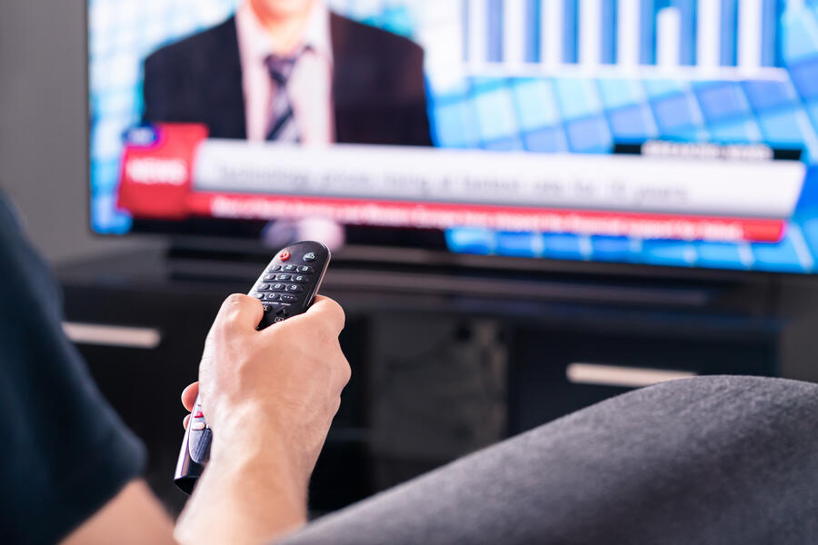 A hand points a remote at a TV. The TV is playing news coverage.