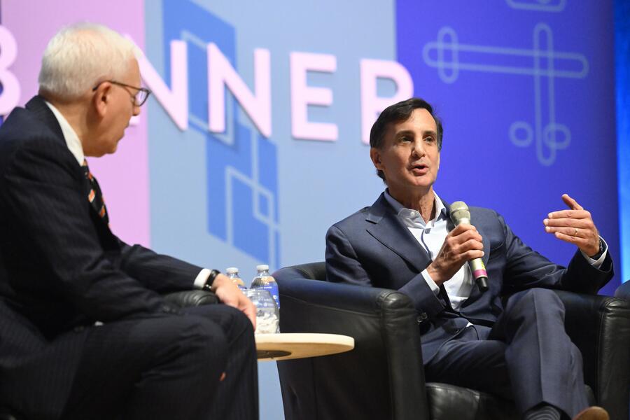 Ron Daniels speaks into a microphone while sitting next to Daniel M. Rubenstein. Both are wearing suits.