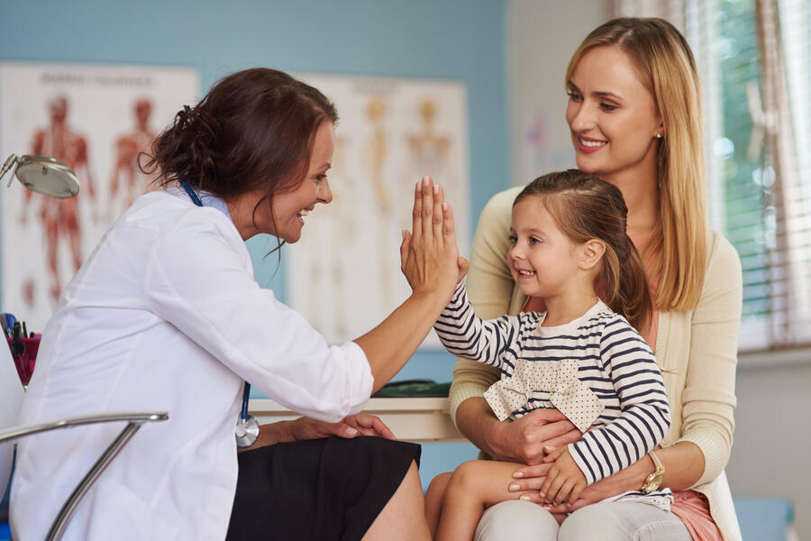 A young girl sitting on her mother's lap in a medical office gives a high five to her pediatrician.