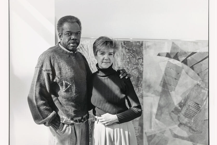 In this black and white photo, two pose together in front of a painting