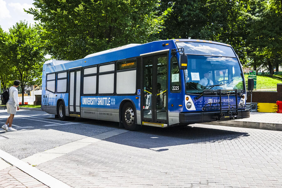 A blue Johns Hopkins University shuttle bus parked at an angle on a sunny day in front of green trees