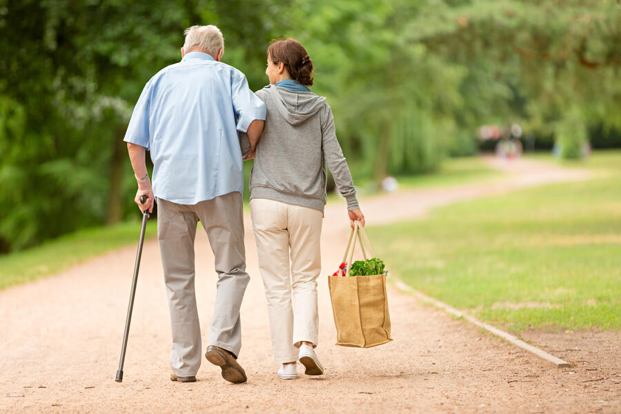 A woman carrying a bag of groceries walks with her elderly father, who uses a cane