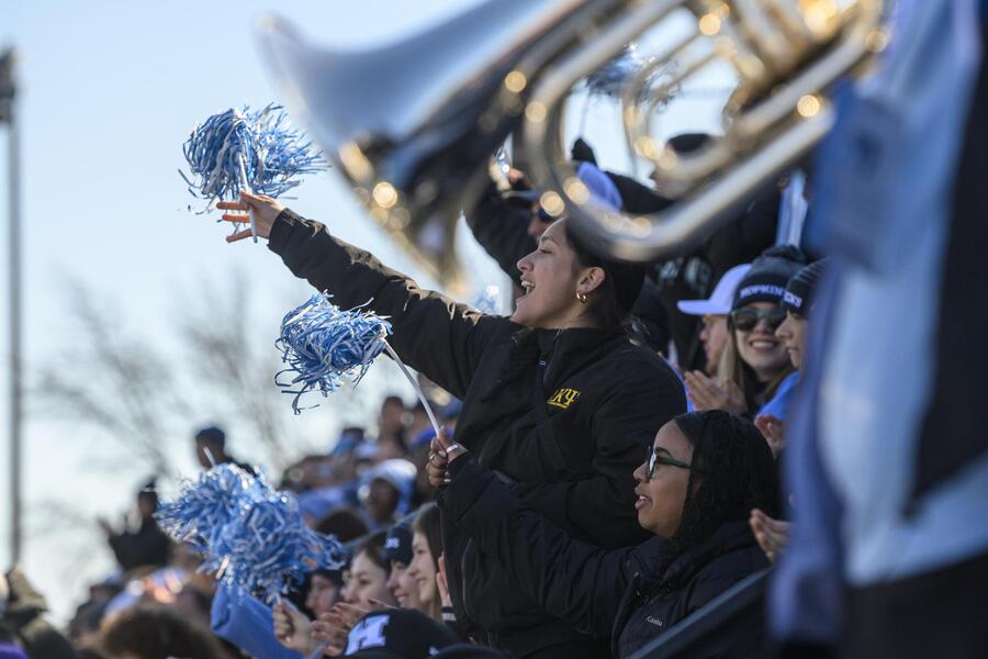 A crowd cheers at a sports game. One attendee is standing up, waving a blue and white pom-pom