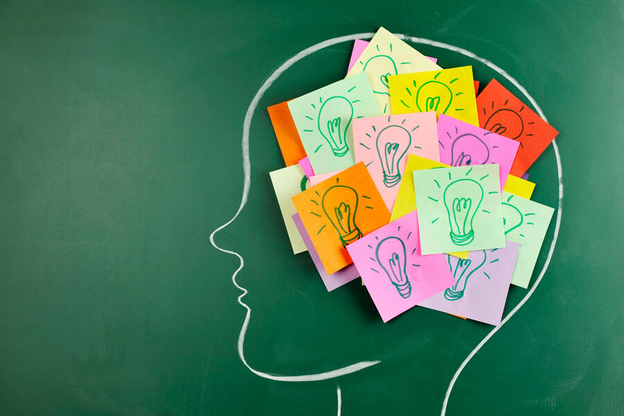 Outline of a head on a green chalkboard that has sticky notes with lightbulbs on the brain