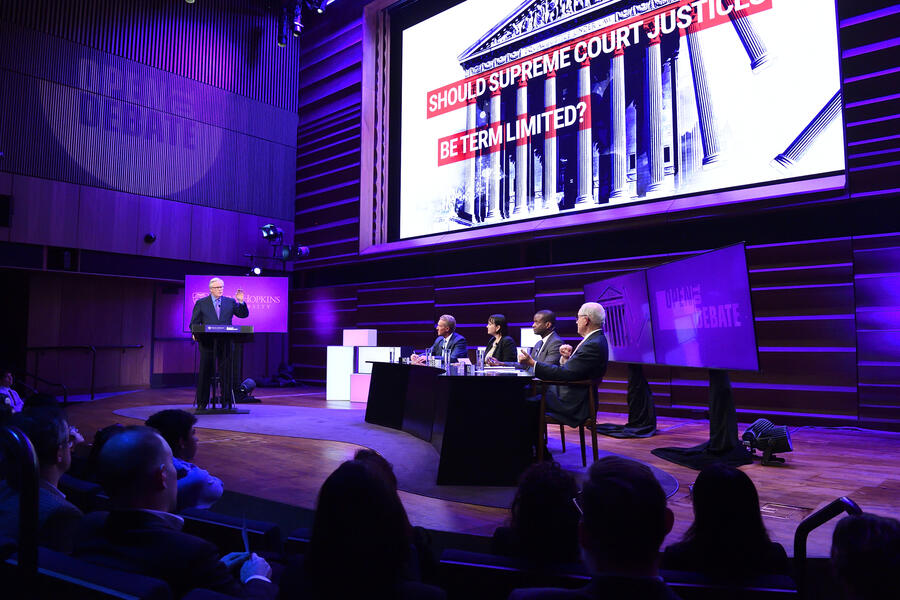 Four adults sit behind a table on-stage. A moderator sits on a stool, asking them a question. Behind them is a screen that reads 