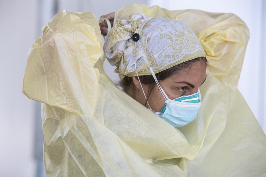 A medical professional in a bandana and medical mask puts on a surgical gown.