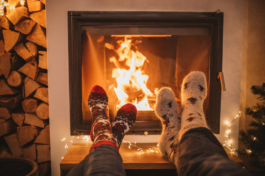 Two pair of crossed feet in cozy socks rest on an ottoman in front of a blazing fire