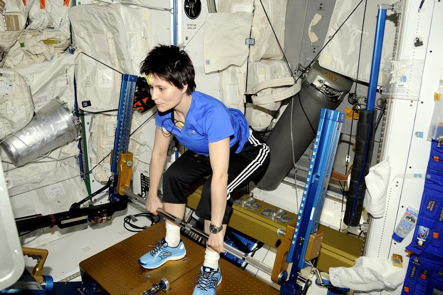 Italian astronaut Samantha Cristoforetti, wearing a blue shirt and black athletic pants, performs a deadlift exercise using the advanced Resistive Exercise Device (aRED) aboard the International Space Station. The aRED machine, equipped with metal bars and vacuum cylinders, provides resistance training to help astronauts maintain muscle and bone strength in microgravity. The surrounding area is filled with white padded walls, equipment, and storage bags, characteristic of the ISS interior.